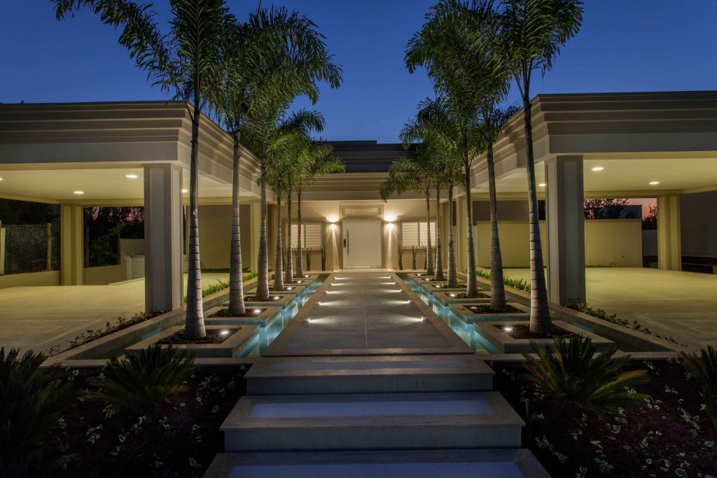 Luxury residence at dusk with palm-lined approach and layered exterior lighting