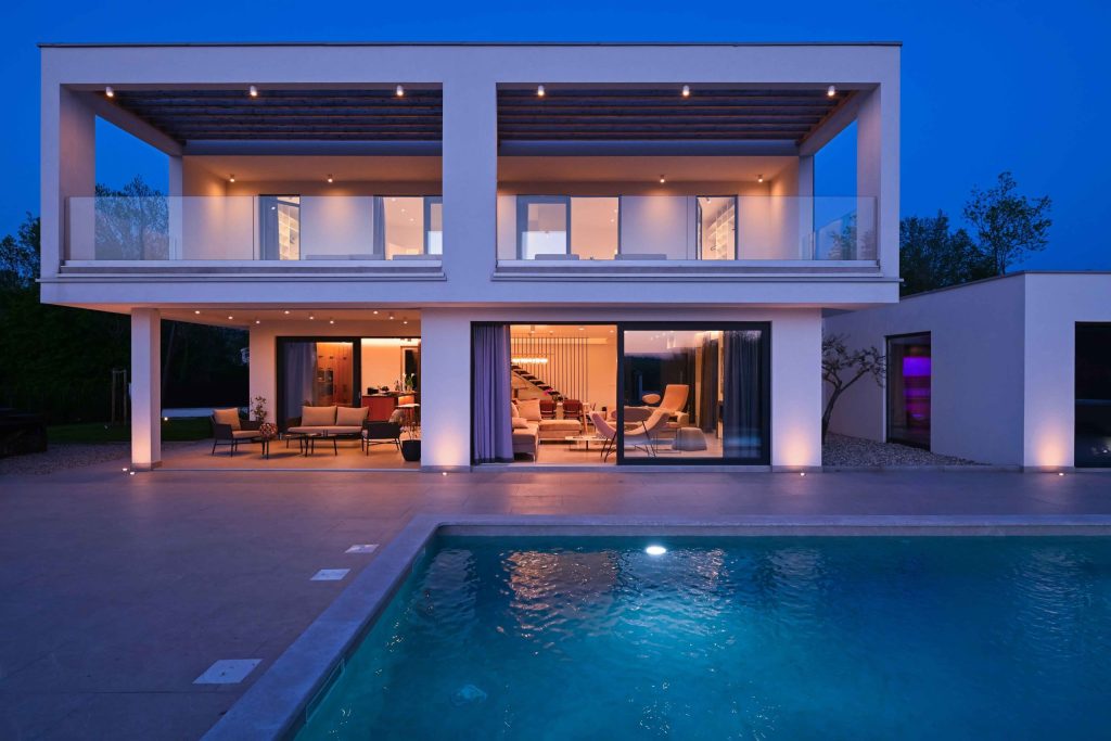 Modern two-story luxury home illuminated at dusk, viewed from a poolside terrace