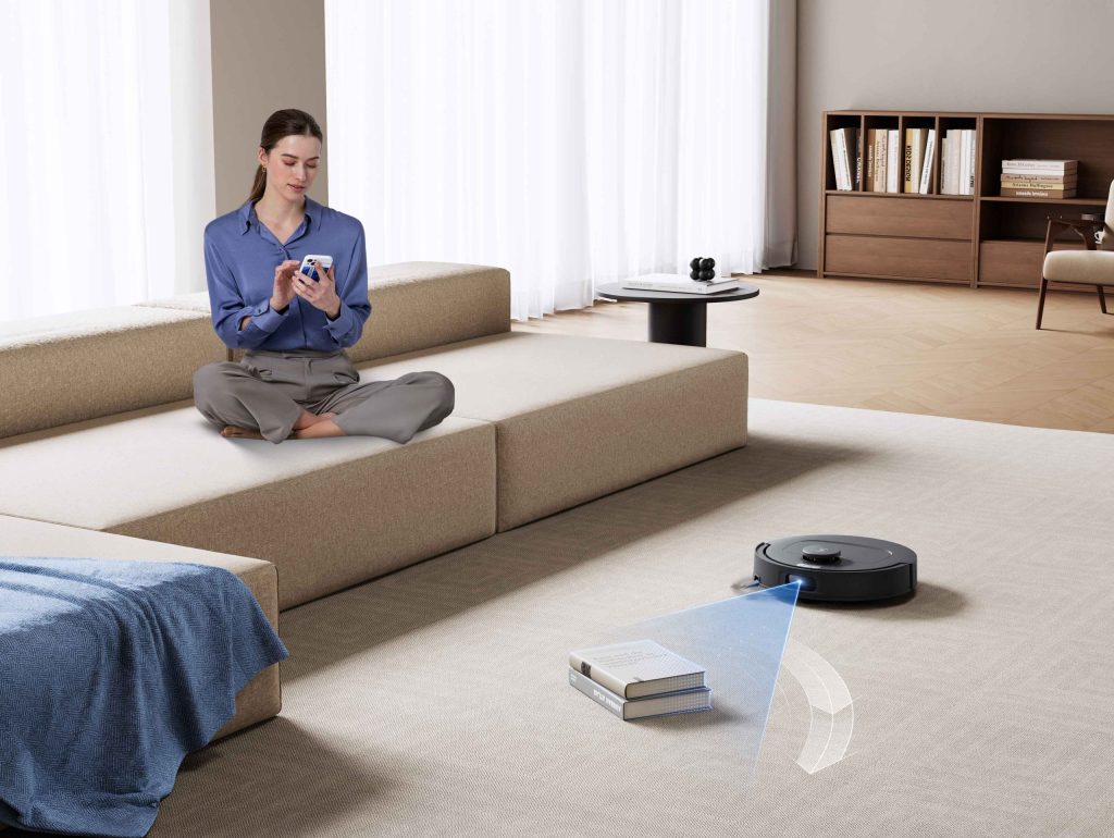 床に置かれた本を回避しながら清掃するDEEBOT N50 OMNIとスマートフォンを操作する人物