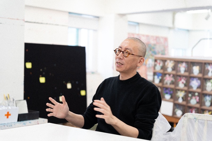 Contemporary artist speaking in a bright studio surrounded by paintings and art materials