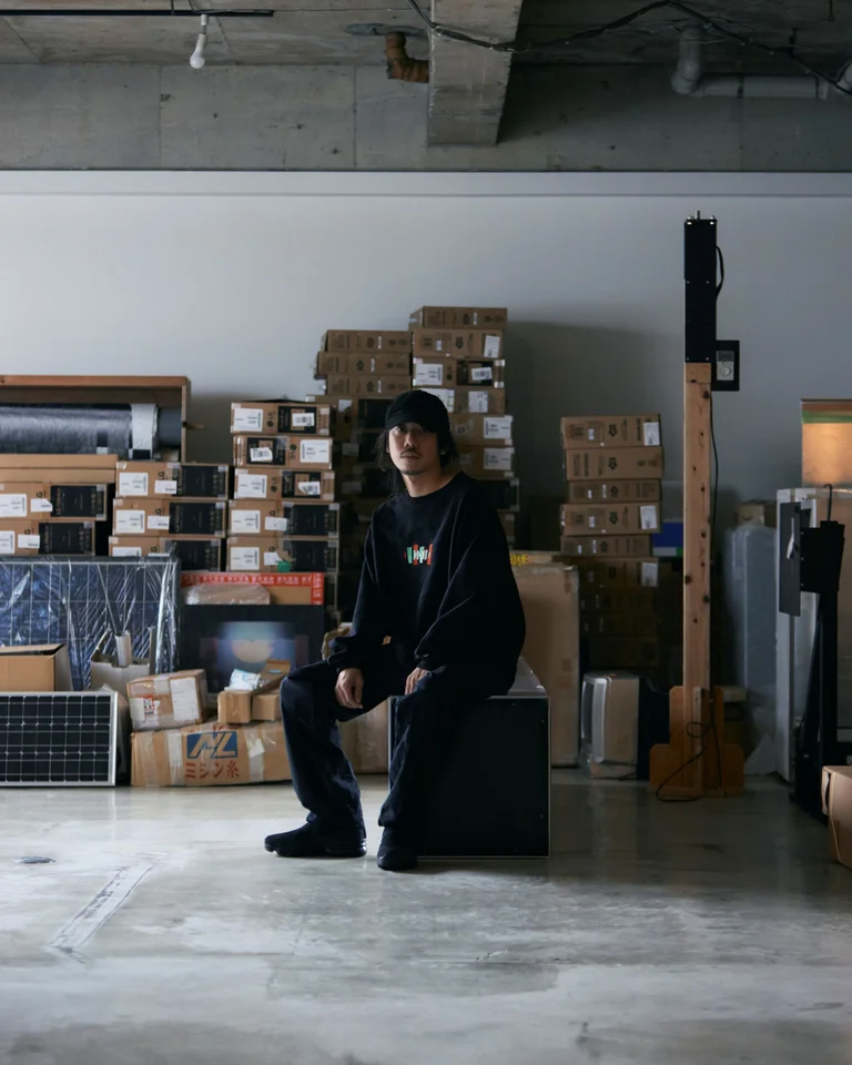 コンクリートの床と積み重ねられた資材を背景に、黒い服を着た人物が座るポートレート