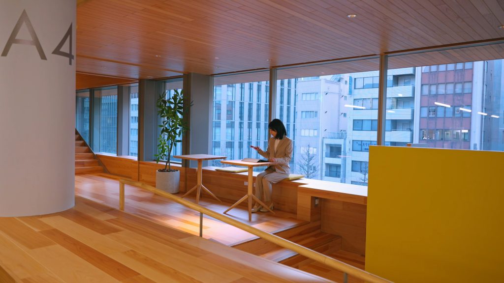Daylit timber-lined perimeter workspace with a person using a smartphone beside large office windows